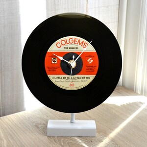 May include: A black vinyl record clock featuring the text "COLGEMS THE MONKEES" and "A LITTLE BIT ME, A LITTLE BIT YOU." The clock has white hands and is mounted on a white stand. The record label is orange and white.