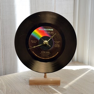 May include: A black vinyl record clock featuring the album title "Goodbye Yellow Brick Road" by Elton John. The clock has gold-colored hands and is mounted on a small wooden stand. The record label has a rainbow design.