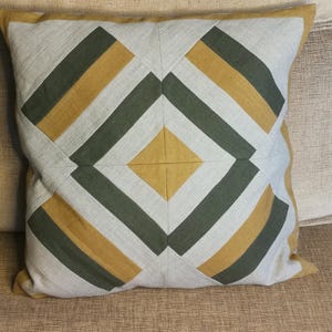 Puede incluir: Cojín decorativo cuadrado con un patrón geométrico en tonos grises, verde oliva y dorado. El cojín está colocado sobre un sofá marrón, mostrando su diseño y textura. El diseño del cojín presenta una forma de diamante central.