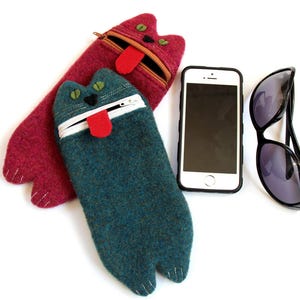 Puede incluir: Dos estuches de fieltro con forma de gato, uno rojo y otro azul verdoso, con ojos negros y una lengua roja que sobresale. Tambi&eacute;n se ve un tel&eacute;fono inteligente negro y unas gafas de sol en la imagen.