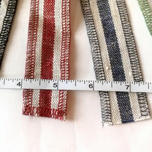 May include: Four different striped fabric ribbons with white and colored stripes. The ribbons are lined up horizontally and measured with a ruler. The ribbons are black and white, red and white, blue and white, and green and white.
