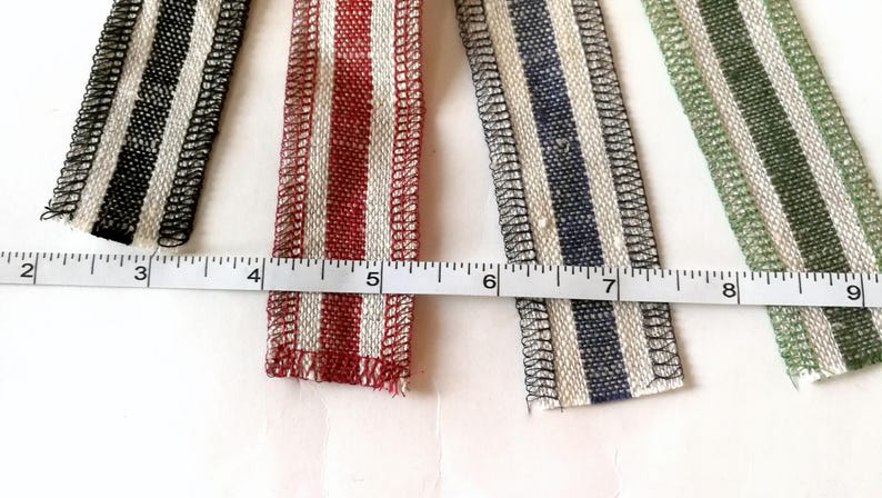 May include: Four different striped fabric ribbons with white and colored stripes. The ribbons are lined up horizontally and measured with a ruler. The ribbons are black and white, red and white, blue and white, and green and white.