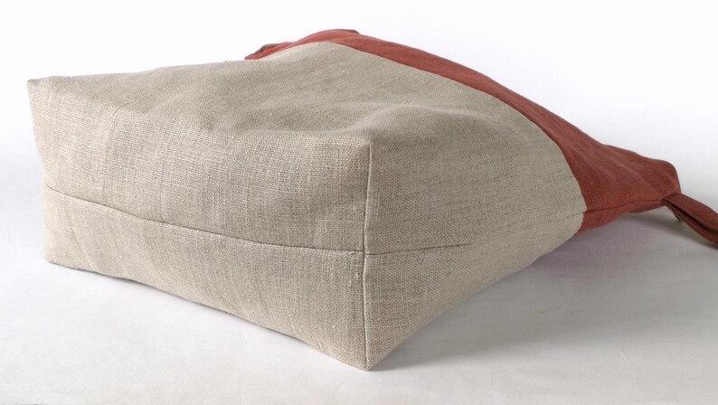 May include: A beige linen tote bag with a red linen panel on the top. The bag is shown from the bottom, with the seams visible.
