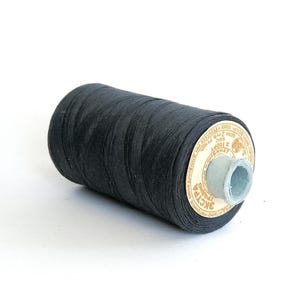 May include: A spool of black thread, tightly wound, with a paper label featuring text and a light blue core. The thread is a deep, solid black, and the spool is set against a plain white background.