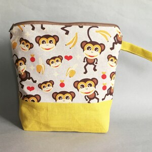 Può includere: Una pochette con cerniera gialla e bianca con un tiretto della cerniera marrone. La pochette è realizzata in tessuto e presenta un motivo di scimmie, banane e cuori. La pochette ha un manico giallo.