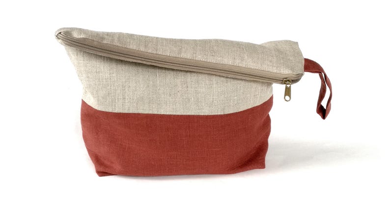 May include: A beige and red linen zippered pouch with a handle. The pouch is made of a natural fabric and has a simple design.