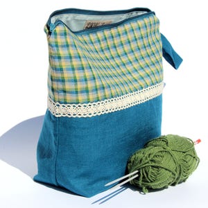 Puede incluir: Una bolsa de hilo de tela a cuadros verde azulado y amarillo con un ribete de encaje blanco. La bolsa está abierta, revelando un interior azul claro. Dos ovillos de hilo verde y agujas de tejer están junto a la bolsa.