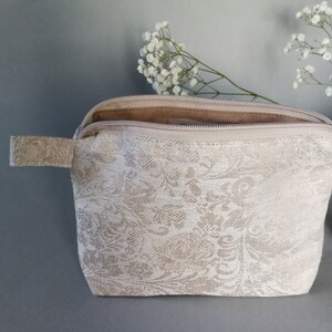 May include: A beige zippered pouch with a white floral pattern. The pouch is open and the zipper is visible. The pouch is sitting on a gray surface.