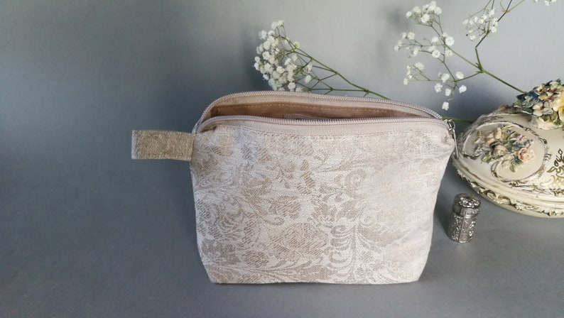 May include: A beige zippered pouch with a white floral pattern. The pouch is open and the zipper is visible. The pouch is sitting on a gray surface.