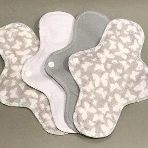 Puede incluir: Cuatro compresas menstruales de tela reutilizables con estampado de mariposas. Dos compresas son grises con mariposas blancas y dos compresas son blancas con mariposas grises.