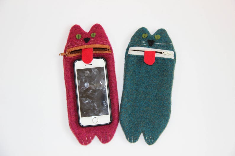 Puede incluir: Dos fundas para tel&eacute;fono de fieltro con forma de gato, una roja y otra azul turquesa, con una cremallera y una lengua de fieltro que sobresale. Ambas fundas tienen un tel&eacute;fono blanco en su interior.