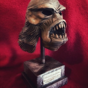 May include: A detailed sculpture of a creature's head, with sharp teeth and a menacing expression. The sculpture is mounted on a dark, wooden-like base with a metal rod. The background is a deep red fabric.