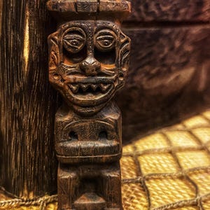 May include: A carved wooden tiki statue with a smiling face, detailed features, and a leather strap. The dark brown statue has a hat and is set against a textured background.