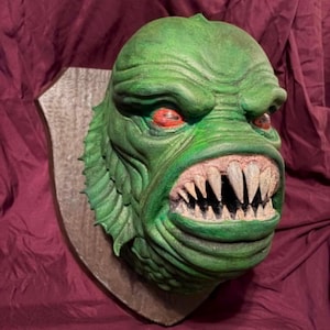 May include: A green monster head with sharp teeth and red eyes, mounted on a wooden plaque. The creature has a textured, scaly appearance and a menacing expression. The background is a deep burgundy fabric.