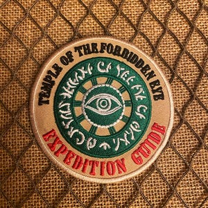 May include: A tan and red embroidered patch with the text "Temple of the Forbidden Eye Expedition Guild" and a green eye with a white circle around it.