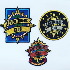 Könnte beinhalten: Drei gestickte Aufnäher mit einem Kompass-Design und dem Text "Adventurers Club". Der obere Aufnäher ist blau und gelb, der mittlere Aufnäher ist schwarz und gelb, und der untere Aufnäher ist schwarz, rot und gelb. Der untere Aufnäher enthält auch den Text "Member".