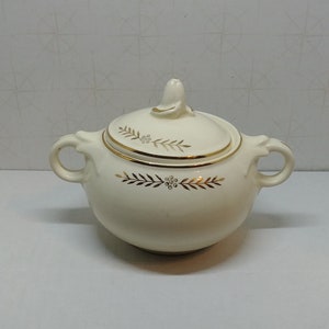 May include: Cream-colored ceramic sugar bowl with a lid and two handles. The lid has a small finial and is decorated with a gold band and gold leaf pattern. The bowl also has a gold leaf pattern.