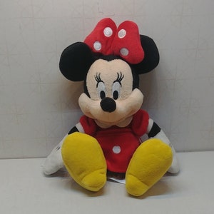 Puede incluir: Una mu&ntilde;eca de peluche de Minnie Mouse. La mu&ntilde;eca tiene orejas negras, un lazo rojo con lunares blancos y un vestido rojo. Se ven sus pies amarillos y tiene guantes blancos. La mu&ntilde;eca est&aacute; sentada.