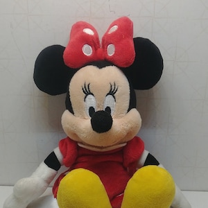 Pu&ograve; includere: Una bambola di peluche di Minnie Mouse, con un vestito rosso, scarpe gialle e un fiocco rosso a pois bianchi. La bambola ha orecchie nere, un naso nero e guanti bianchi. La bambola &egrave; seduta su una superficie bianca.