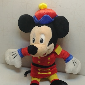 May include: A plush Mickey Mouse toy in a red and yellow uniform with gold accents and a matching hat. The toy has black ears, a white face, and white gloves. The toy is approximately 10 inches tall.