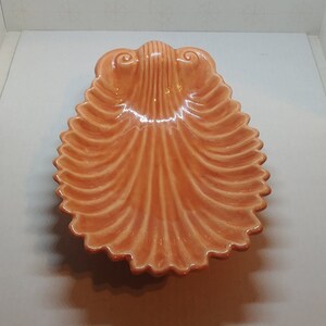 May include: A glossy, peach-colored ceramic dish shaped like a large scallop shell. The dish features a series of raised, radiating ridges that extend from a central hinge area toward the scalloped, wavy edges, creating a textured, fan-like appearance.