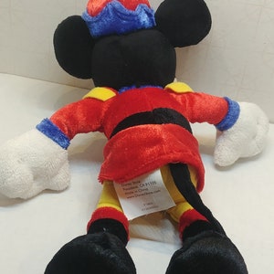 May include: A Mickey Mouse plush toy wearing a red jacket with black trim, yellow accents, and a blue and red hat. The toy has black ears, a black tail, and white gloves and feet. A tag is attached.