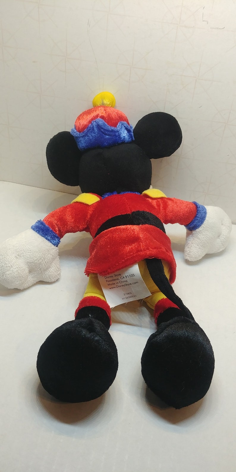 May include: A Mickey Mouse plush toy wearing a red jacket with black trim, yellow accents, and a blue and red hat. The toy has black ears, a black tail, and white gloves and feet. A tag is attached.
