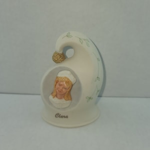 May include: A cream-colored ceramic figurine in the shape of a stylized hat. The figurine features a portrait of a girl named Clara, with blonde hair and a white hat. The hat is decorated with green holly leaves and a gold-colored ball.