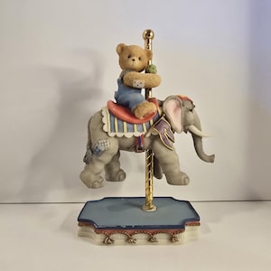 May include: A carousel figurine featuring a teddy bear riding a gray elephant. The bear wears blue overalls and holds a green ball. The elephant has a red saddle and decorative trim. The base is blue with gold accents.