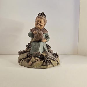 May include: A whimsical figurine of a smiling person in a teal dress and a ruffled collar, holding a brown accordion. The figure is adorned with a pointed hat and sits atop a base of leaves and decorative elements.