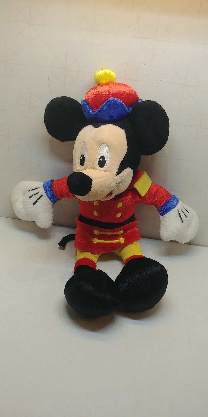 May include: A plush Mickey Mouse toy in a red and yellow uniform with gold accents and a matching hat. The toy has black ears, a white face, and white gloves. The toy is approximately 10 inches tall.