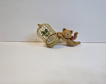 May include: A whimsical figurine of a tan cat with a red bow, appearing to push a gold-colored birdcage. Inside the cage is a green bird. The cage is round with a flat base. The cat has a playful expression, suggesting a decorative item.