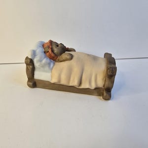 Puede incluir: Figura de cerámica de un oso acostado en una cama. El oso lleva una gorra roja y está cubierto con una manta beige. La cama tiene un marco marrón y una almohada azul.