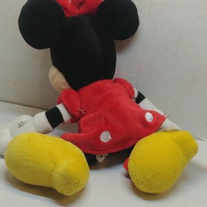 Pu&ograve; includere: Una bambola di peluche di Minnie Mouse, con testa nera, grandi orecchie e fiocco rosso. Indossa un vestito rosso a pois bianchi e scarpe gialle. La bambola &egrave; seduta su una superficie bianca.