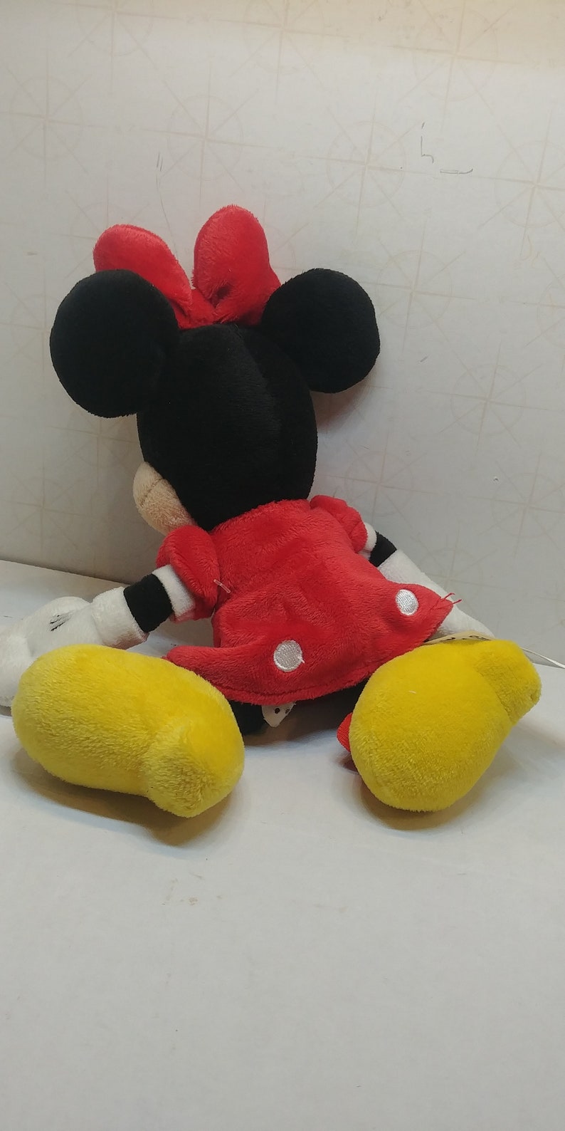 Pu&ograve; includere: Una bambola di peluche di Minnie Mouse, con testa nera, grandi orecchie e fiocco rosso. Indossa un vestito rosso a pois bianchi e scarpe gialle. La bambola &egrave; seduta su una superficie bianca.