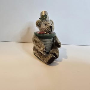 Puede incluir: Figura de oso de peluche hecha a mano con un sombrero de dedal plateado y un cuerpo de carrete verde. El oso tiene pelaje marrón y está sentado. Una pequeña hoja verde de acebo está en el sombrero. El carrete está envuelto con hilo gris.