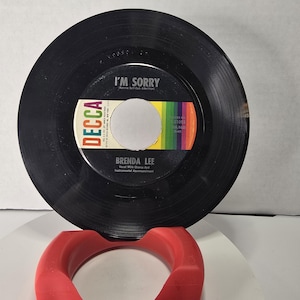 May include: A black 45 RPM vinyl record by Brenda Lee titled I'm Sorry on the Decca label. The center label features a rainbow-colored stripe design, white text, and the Decca logo. The record is held upright by a curved red plastic stand.