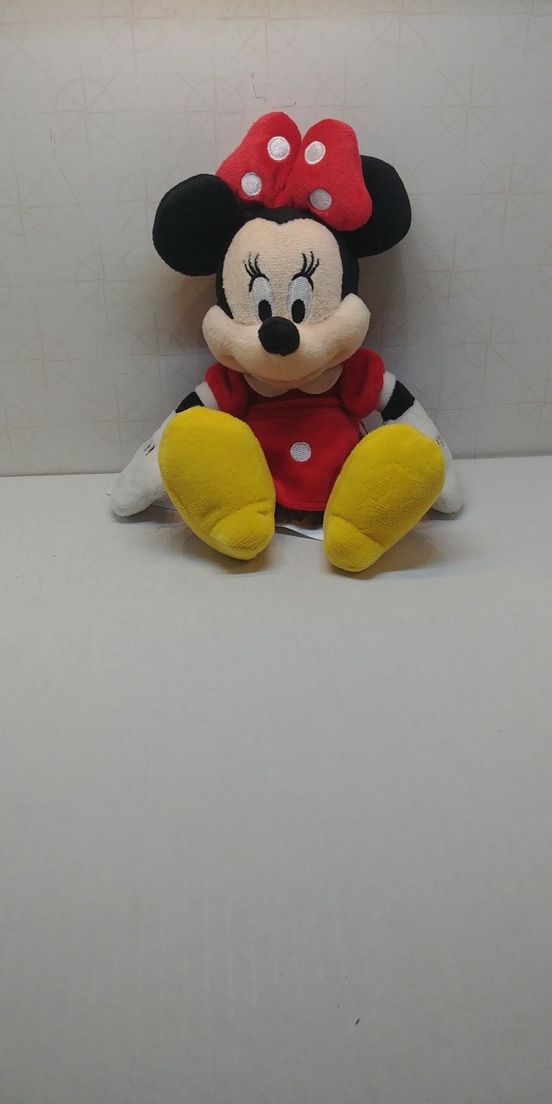 Puede incluir: Una mu&ntilde;eca de peluche de Minnie Mouse. La mu&ntilde;eca tiene orejas negras, un lazo rojo con lunares blancos y un vestido rojo. Se ven sus pies amarillos y tiene guantes blancos. La mu&ntilde;eca est&aacute; sentada.