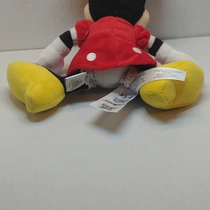 Puede incluir: Un mu&ntilde;eco de peluche de Minnie Mouse, visto desde atr&aacute;s, con orejas negras y un lazo rojo. Lleva un vestido rojo con lunares blancos, guantes blancos y zapatos amarillos. Se ven etiquetas.