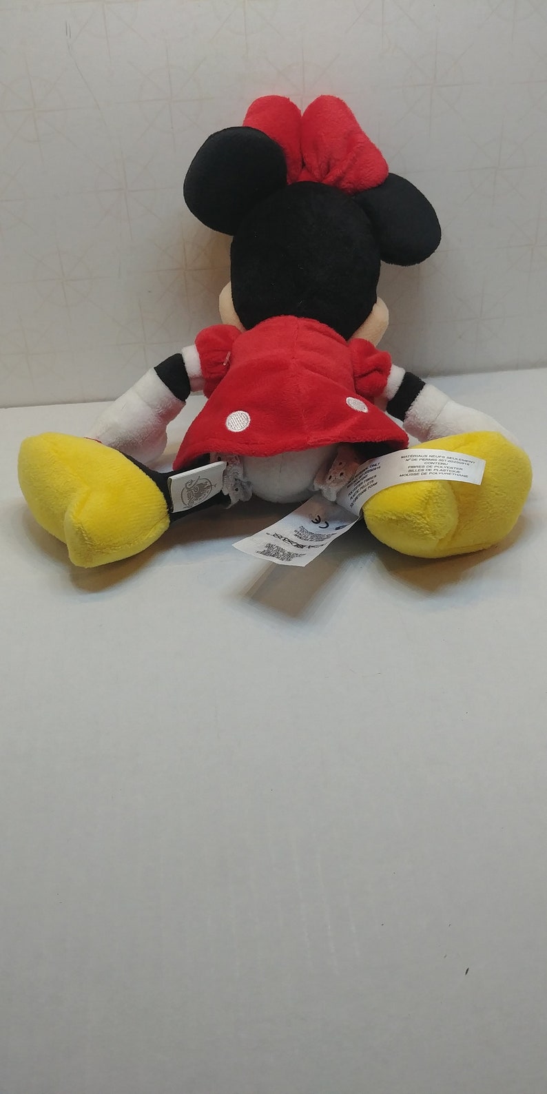 Puede incluir: Un mu&ntilde;eco de peluche de Minnie Mouse, visto desde atr&aacute;s, con orejas negras y un lazo rojo. Lleva un vestido rojo con lunares blancos, guantes blancos y zapatos amarillos. Se ven etiquetas.