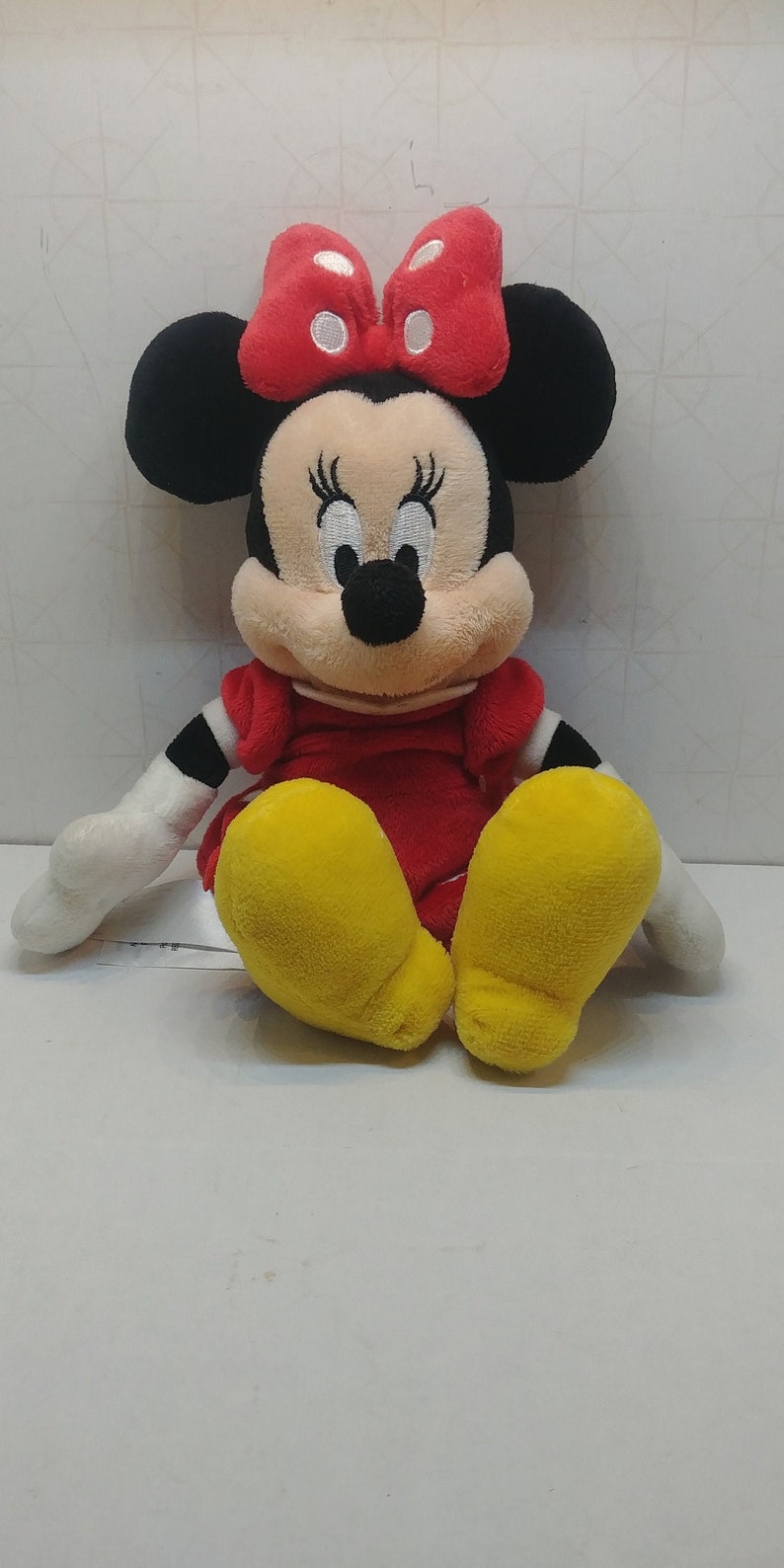 Pu&ograve; includere: Una bambola di peluche di Minnie Mouse, con un vestito rosso, scarpe gialle e un fiocco rosso a pois bianchi. La bambola ha orecchie nere, un naso nero e guanti bianchi. La bambola &egrave; seduta su una superficie bianca.