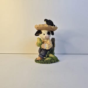 May include: A whimsical figurine of a cow dressed as a farmer, wearing a straw hat with a blackbird perched on top. The cow is holding corn and standing next to a wooden fence. The figurine is painted with vibrant colors and detailed features.