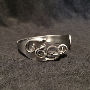 May include: A silver-toned metal cuff bracelet crafted from a repurposed fork. The design features the tines of the fork bent into intricate, swirling spiral patterns that extend outward from the main band, creating a continuous open-ended loop.