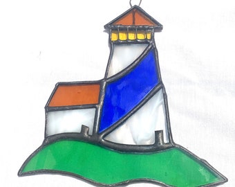 May include: Stained glass lighthouse with a brown roof, white tower, blue accents, and a green base. The lighthouse has a metal frame and a small loop for hanging. A decorative nautical item.