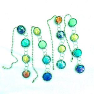 Puede incluir: Tres cadenas plateadas con cuentas redondas de vidrio de colores. Las cuentas son en tonos de azul, verde, amarillo y naranja. Las cadenas están conectadas entre sí por pequeños anillos metálicos.