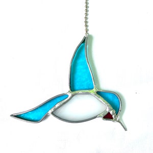 Puede incluir: Un adorno de colibrí de vidrio emplomado con cuerpo azul y pico rojo, colgado de una cadena plateada.