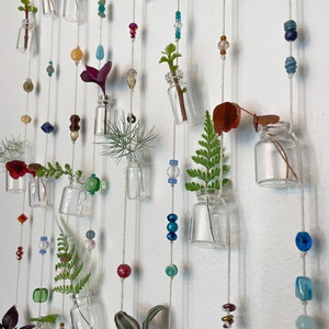 May include: A wall hanging with multiple small glass bottles filled with greenery and colorful beads. The bottles are suspended from a white wall by strings with beads in various colors, including blue, green, red, and orange.