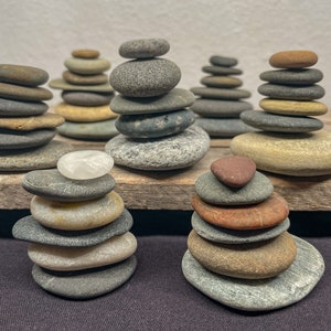May include: A collection of balanced stone stacks, each composed of smooth, flat rocks in various sizes and colors, including gray, brown, and tan. The stacks are arranged on a wooden surface, creating a natural and serene aesthetic. The image showcases a zen-like theme.