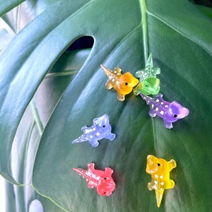 May include: A collection of colorful, miniature dinosaur figurines in shades of yellow, orange, green, purple, blue, and red. Each dinosaur has white polka dots. The figurines are displayed on a large, green leaf.