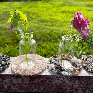 May include: Four small glass vases with different colored flowers are arranged on a wooden surface. Each vase is surrounded by a ring of small stones.